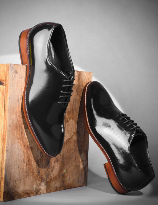 Pair of black dress shoes with brown soles on a wooden block against a gray background