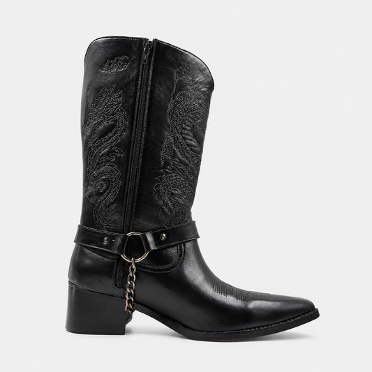 A black leather cowboy boot with a pointed toe, block heel, decorative embossed western pattern on the shaft, and a side strap with a ring and chain detail, shown against a light gray background.