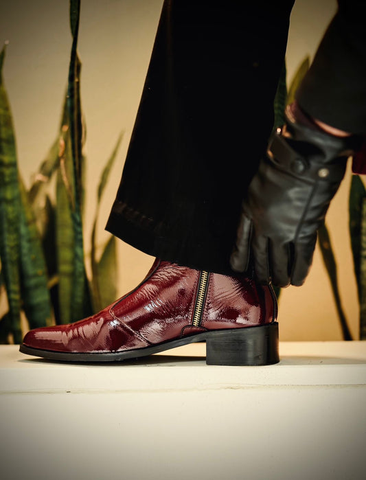 A close-up of a stylish cherry red pointed-toe boot in glossy patent leather with a side zip closure, worn by a person dressed in black trousers and leather gloves. The boot is elevated on a white platform with green snake plants in the background, creating a bold and luxurious contrast.
