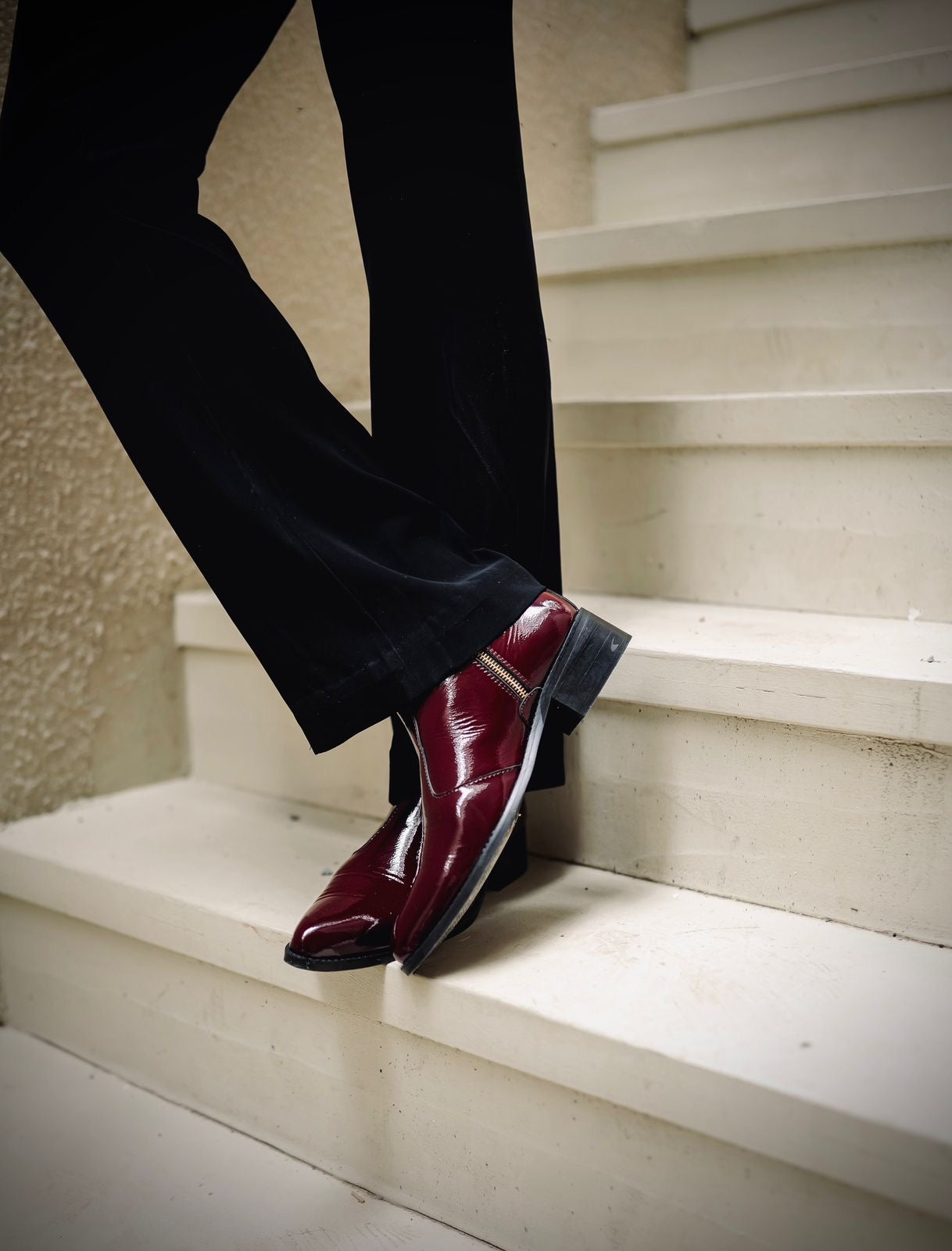 Men’s luxury cherry red pointed-toe Chelsea boots in patent leather with side zip closure, worn with black trousers and leather gloves; sleek and stylish footwear ideal for formal and high-fashion outfits.