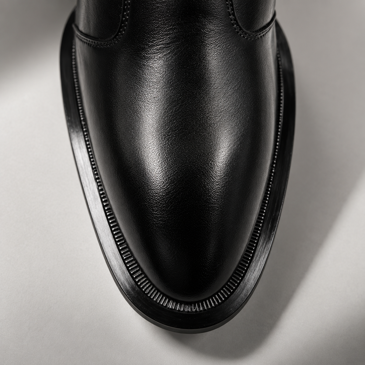 Close-up of a black leather boot toe showing the smooth leather texture, decorative stitching, and a thick ridged black sole on a light gray background.